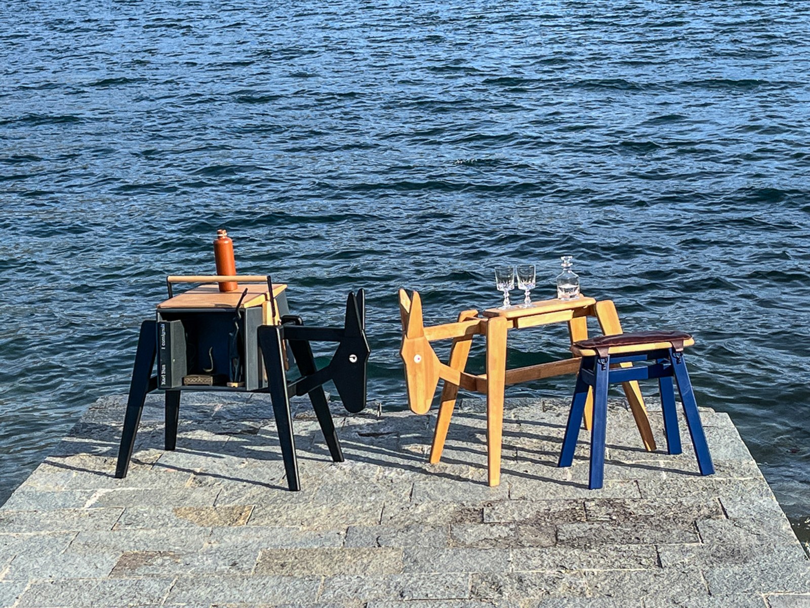 Donkey by NOEMAR — a black side table, a natural-wood frame, and a blue stool arranged on a stone quay by the water, set with glasses and a decanter.
