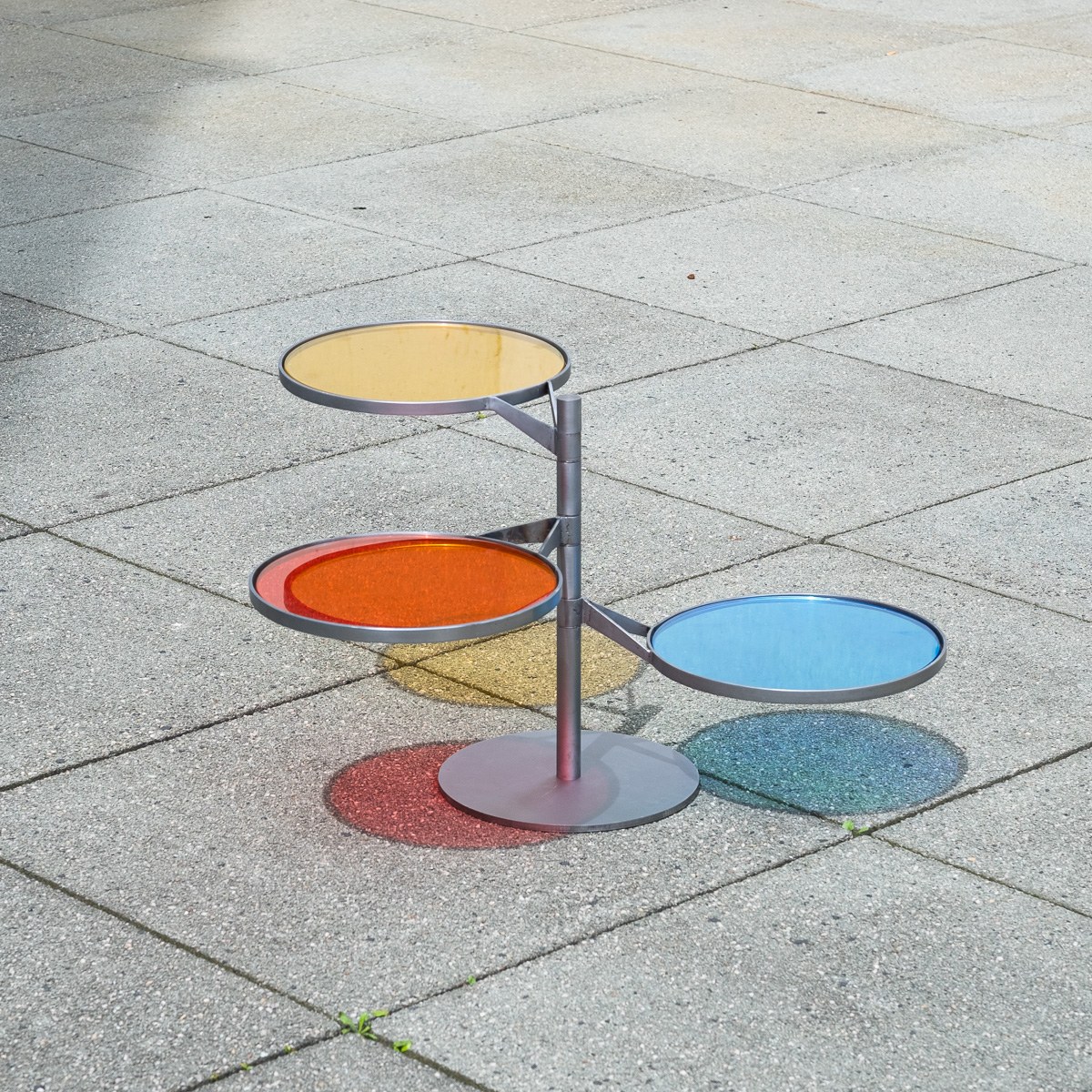 FLTR by NOEMAR — a rotatable steel side table with three tinted glass discs in yellow, red, and blue, casting overlapping colored light onto the ground.