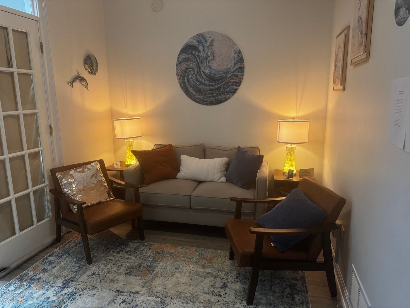 Office seating area with a cream loveseat between two warm table lamps, two brown leather-and-wood armchairs, and a round wave-mosaic artwork on the wall.
