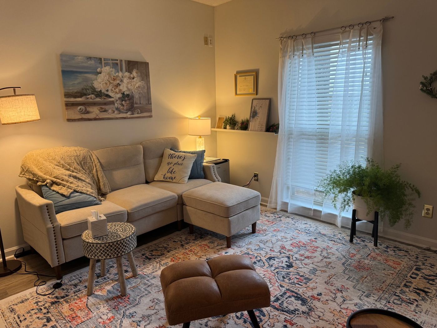 A cream sectional with a knit throw blanket and an ottoman, beside a sheer-curtained window and a potted fern, with a framed license on the wall.