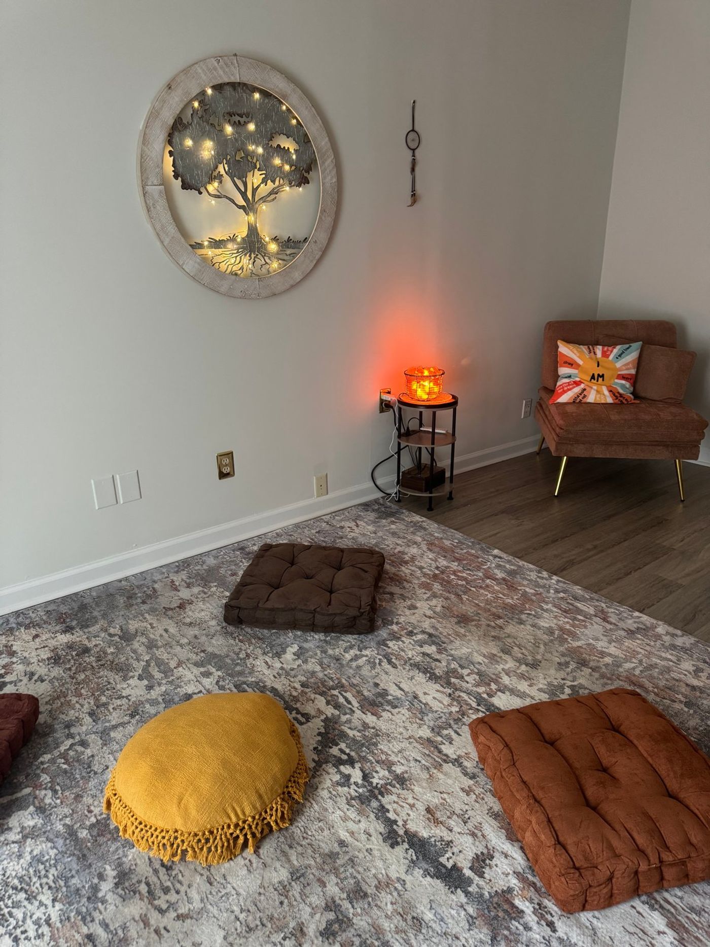 Mustard, brown, and rust floor cushions arranged in a circle on a soft grey rug, below a lit tree-of-life wall piece, with a salt lamp glowing on a side table.