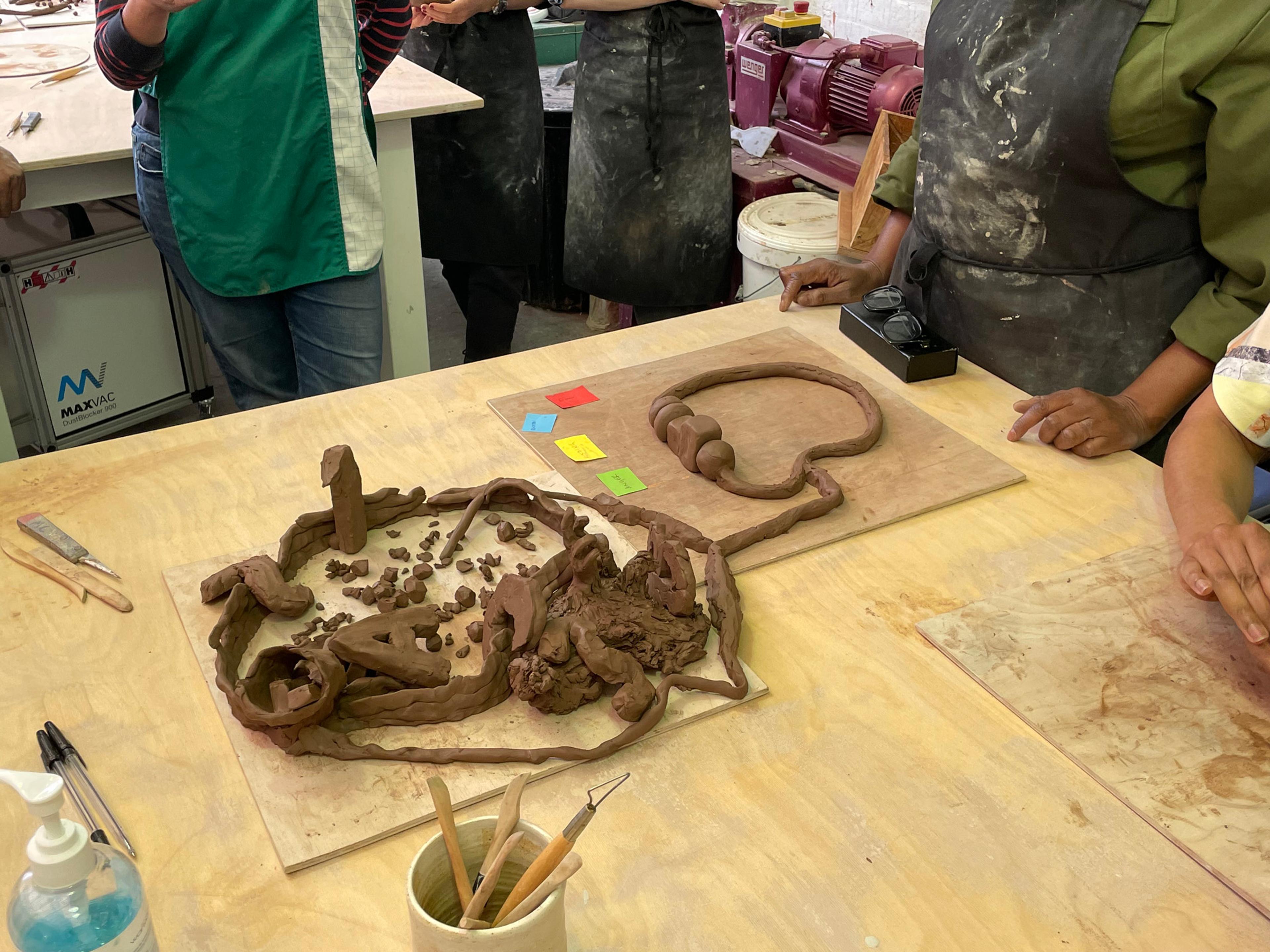 Five people, heads out of picture, stand around a table ooking at some pieces of clay formed into primitive shapes