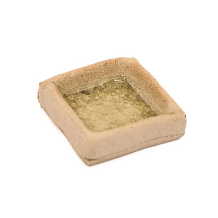 The test tile from a low angle, the raised clay walls and the shallow pool of pale olive glaze crazed across its floor.