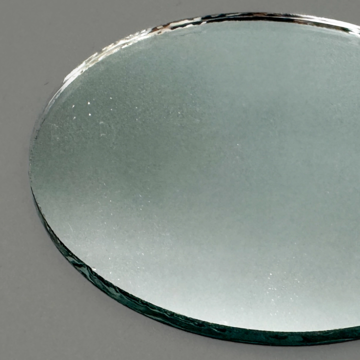 The mirror seen close from above, its silvered face reflecting a soft grey field with the bevelled, green-tinged edge running along one side.