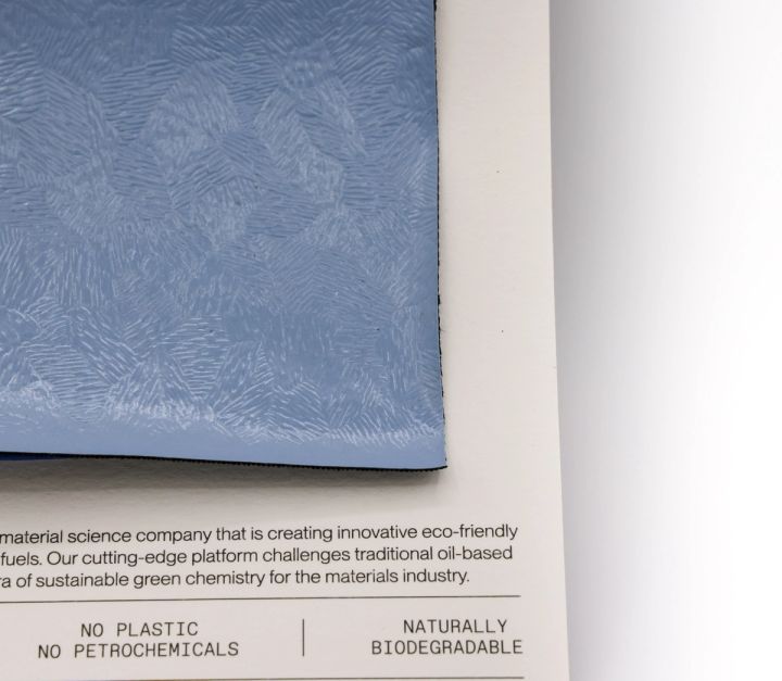 The swatch at an angle across its specification card, the embossed surface catching light, the card reading “no plastic, no petrochemicals, naturally biodegradable”.
