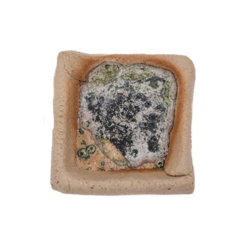 A small square test tile of buff stoneware with raised edges forming a shallow well, holding a fired pool of crusted white and charcoal material rimmed in olive green, its surface bubbled and matte.