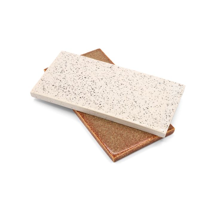 The two tiles stacked at an angle, the speckled cream one lying across the coppery glazed one so both faces and their thin edges show.