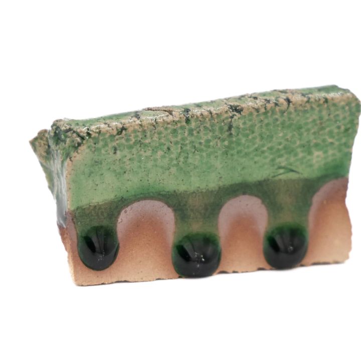A test tile seen edge-on, the green glaze running thickly over the scalloped lower edge and pooling into dark drips beneath.