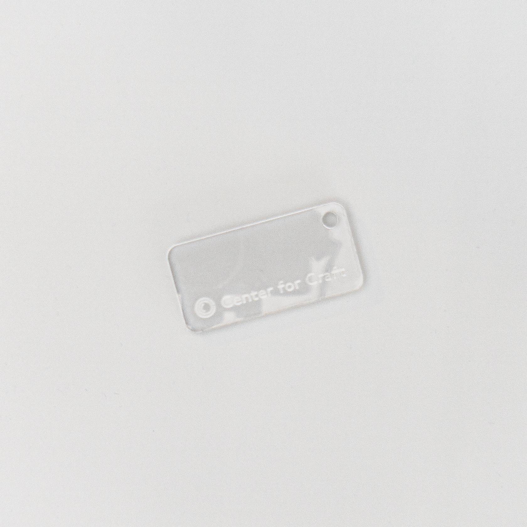 A small clear acrylic rectangle with rounded corners and a hole punched at one end, etched with the Center for Craft name and logo; the material is transparent enough to be barely visible.