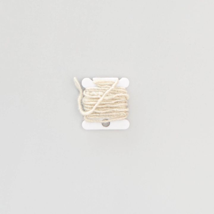 The bobbin seen from further back, the cream yarn wound in even turns with a single strand crossing it diagonally.