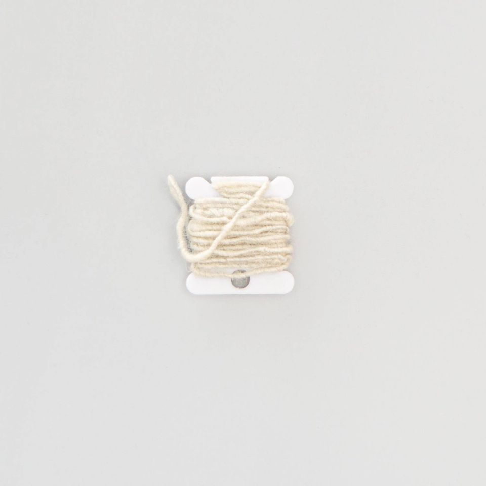 The bobbin seen from further back, the cream yarn wound in even turns with a single strand crossing it diagonally.