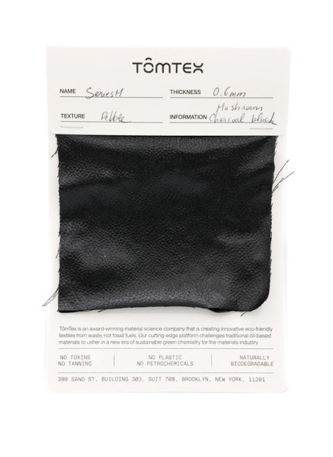 A black leather-like swatch with a fine pebbled grain, stapled to a TômTex specification card recording Series M, 0.6 mm thickness, a pebble texture and mushroom charcoal black.