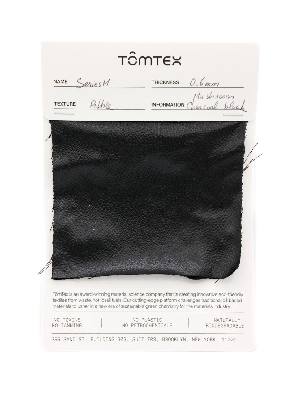 A black leather-like swatch with a fine pebbled grain, stapled to a TômTex specification card recording Series M, 0.6 mm thickness, a pebble texture and mushroom charcoal black.