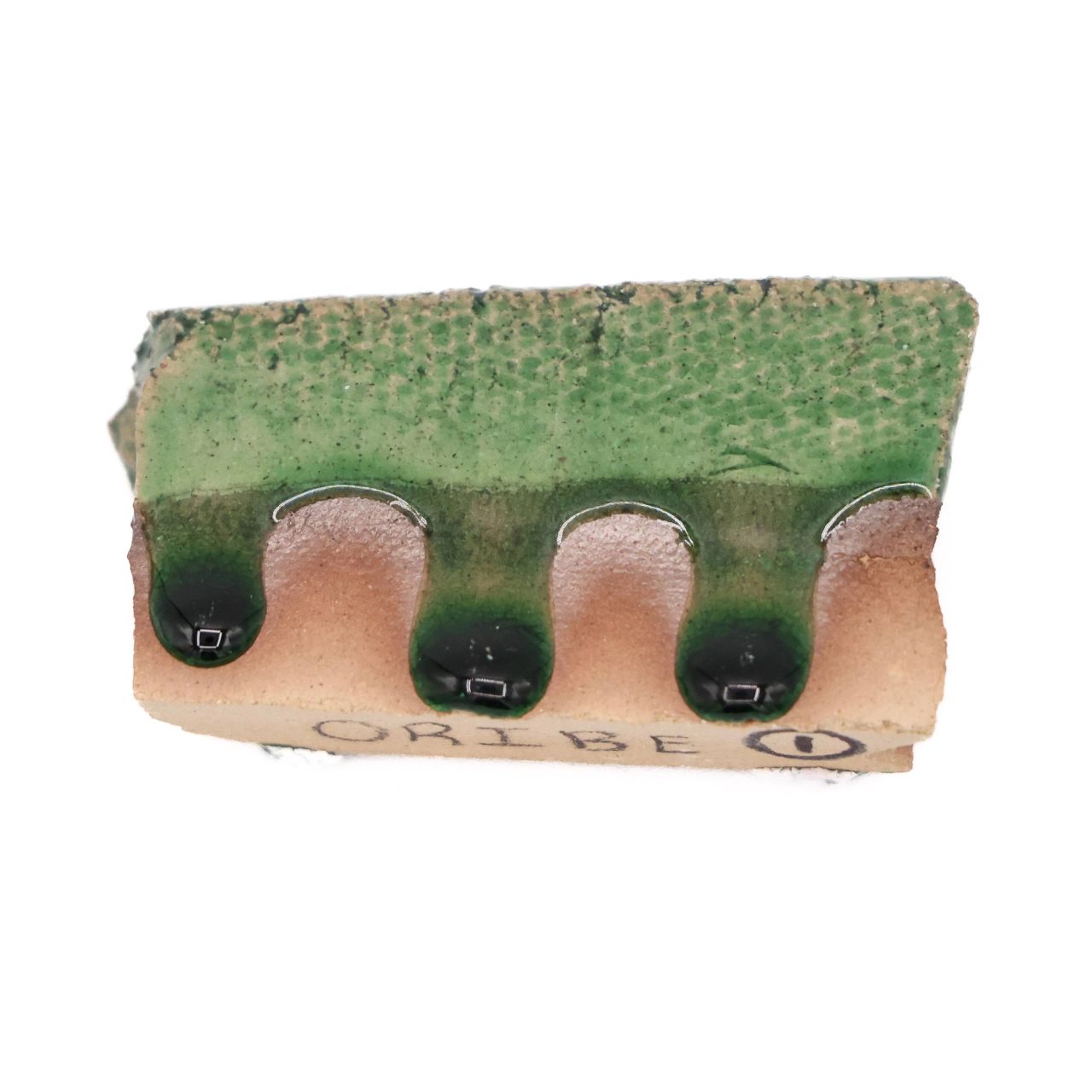 A rectangular ceramic test tile with three rounded scallops along its lower edge; a mottled green glaze covers the upper face and runs down into dark pooled drips, with “ORIBE” pencilled on the pale unglazed clay below.