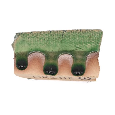 A rectangular ceramic test tile with three rounded scallops along its lower edge; a mottled green glaze covers the upper face and runs down into dark pooled drips, with “ORIBE” pencilled on the pale unglazed clay below.