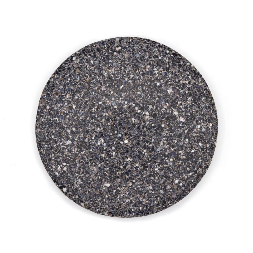 A round tile of dense composite, charcoal grey shot through with pale flecks of crushed shell in white, blue and brown.