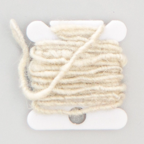 Thick undyed cream wool yarn wound loosely on a white plastic bobbin, one strand trailing across the front, a grey metallic core showing through where the wool thins.
