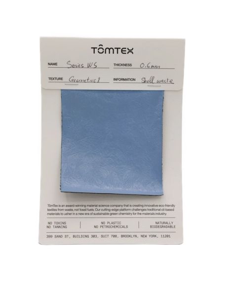 A blue-grey textile swatch with a faint embossed pattern, stapled to a TômTex specification card recording Series WS, 0.6 mm thickness, a geometric texture and shell waste as its source.