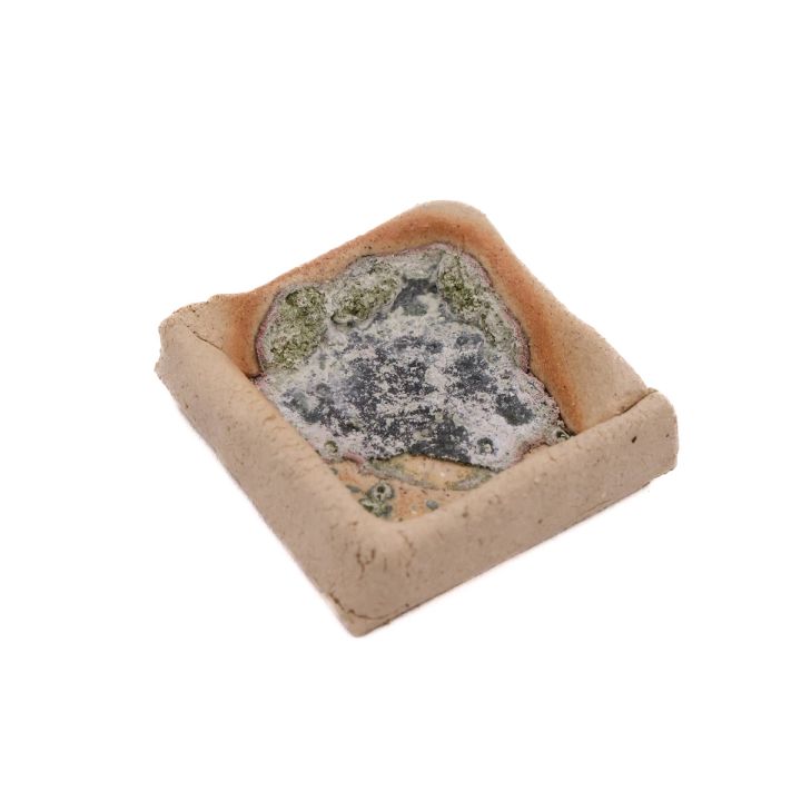 The same test tile seen from the side, its raised clay walls and the depth of the crusted glaze pool now visible.