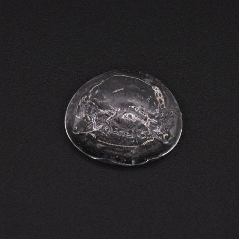 A small clear disc of glass with a rippled, bubbled surface and a few flecks of colour caught inside.
