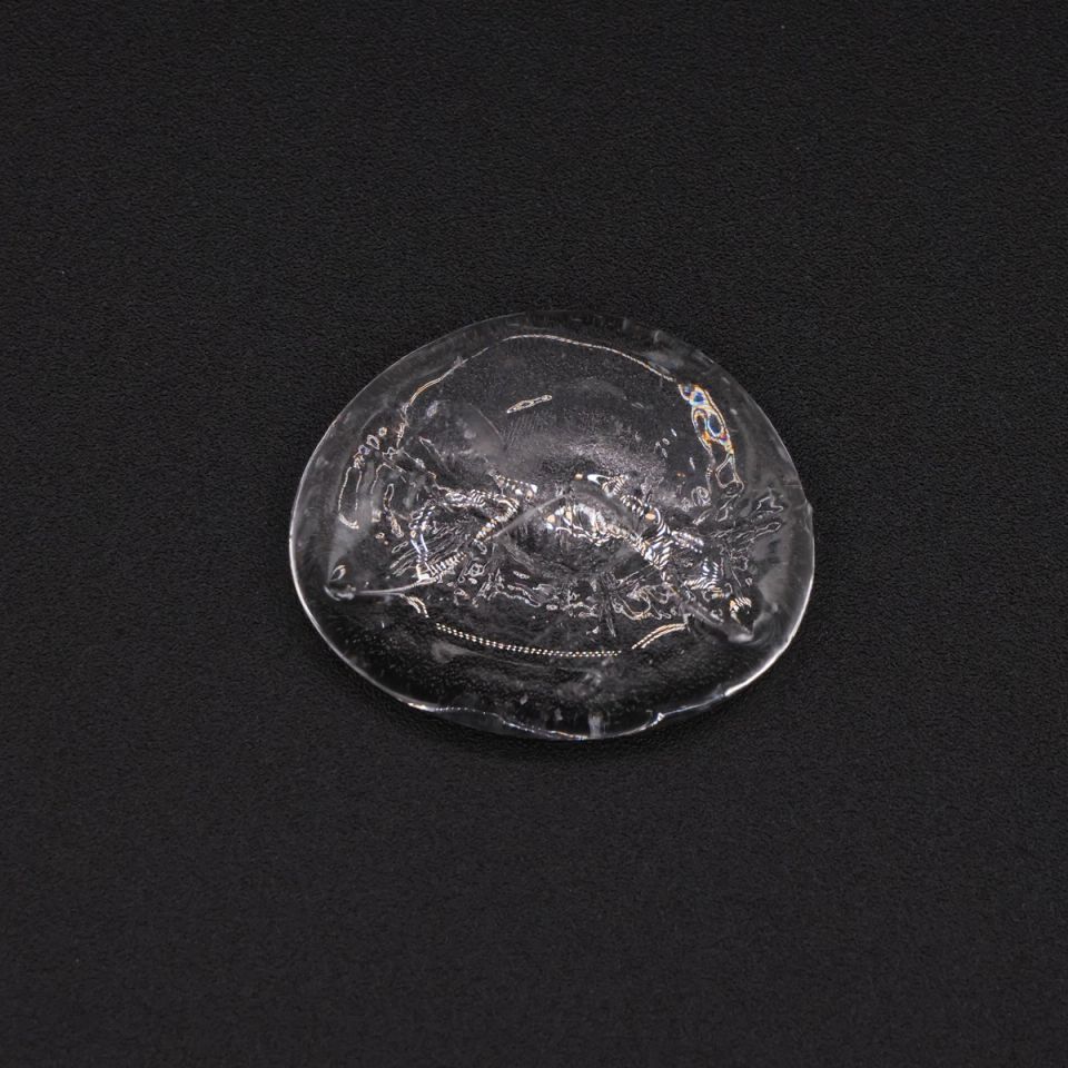 A small clear disc of glass with a rippled, bubbled surface and a few flecks of colour caught inside.