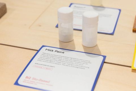 Two small white bottles standing on a pale wood surface behind a printed exhibition card headed “Milk Paint”.