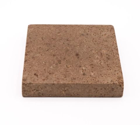 A thick square tile in warm brown seen at an angle to show its depth, the surface densely pitted and flecked with paler particles.