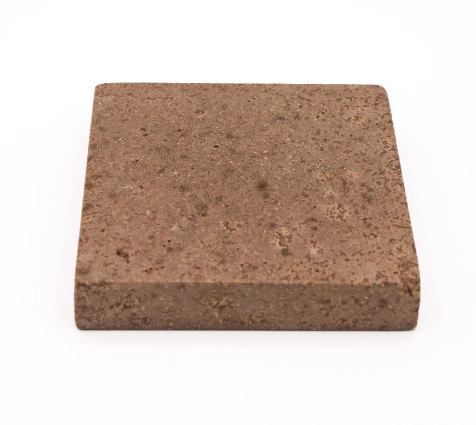A thick square tile in warm brown seen at an angle to show its depth, the surface densely pitted and flecked with paler particles.