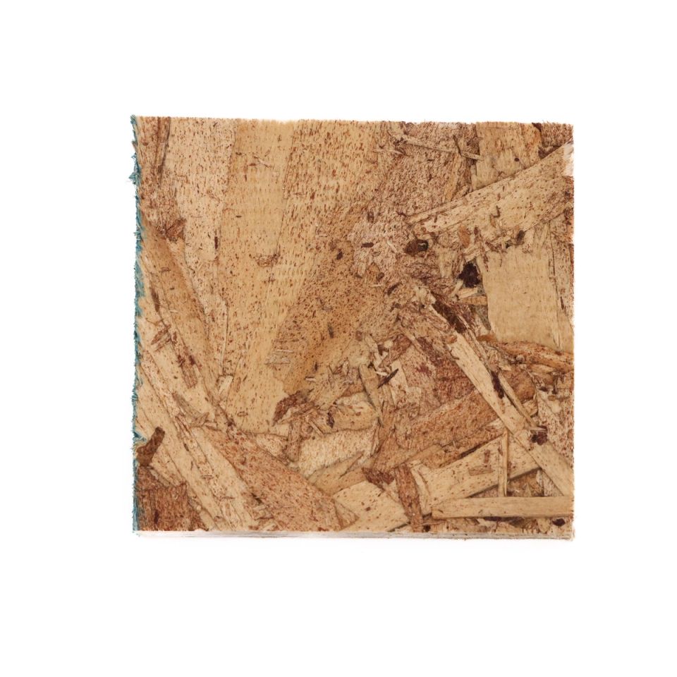 A square of oriented strand board, its face a dense mosaic of flat wood chips in tan and brown pressed at every angle, with a thin teal line along one edge.