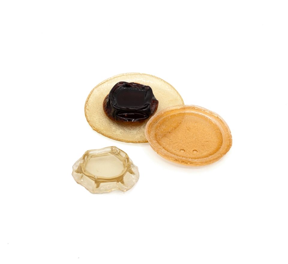 Three cast bioglass pieces in amber and honey tones: a thin translucent oval, a thicker disc with a raised rim, and a smaller faceted block, with a dark caramel lump resting on the largest.