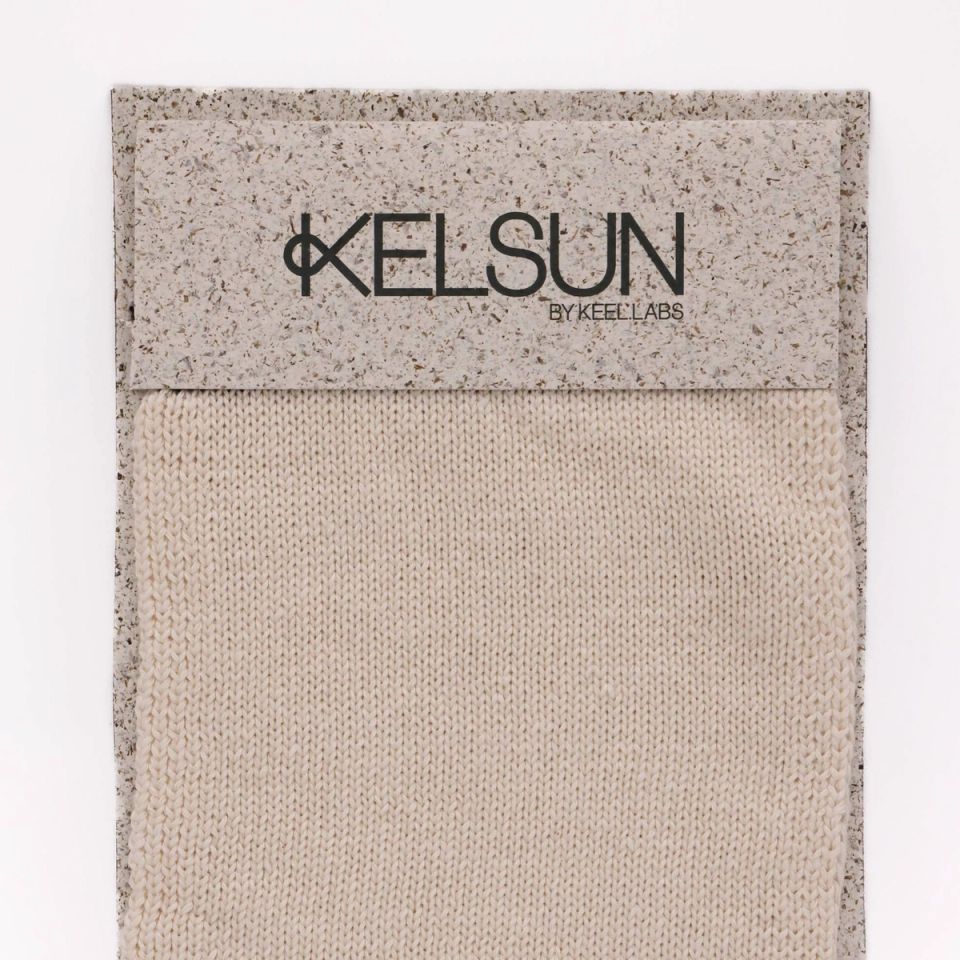 A knitted swatch in undyed cream, stocking stitch running vertically, mounted on a speckled grey board printed “KELSUN by Keel Labs”.