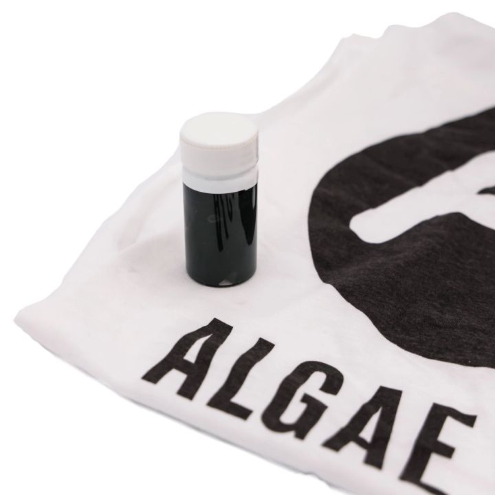 The vial of ink standing on a white T-shirt screen-printed in heavy black type reading “ALGAE”, showing the ink as a finished print.