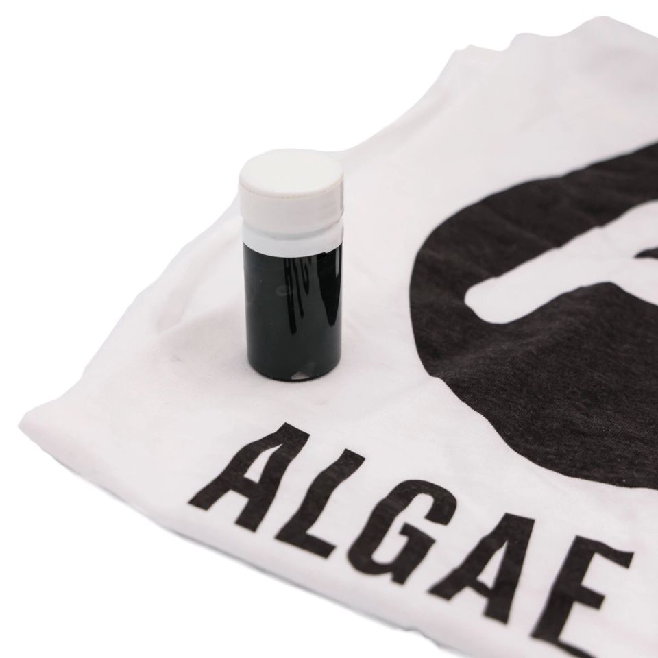 The vial of ink standing on a white T-shirt screen-printed in heavy black type reading “ALGAE”, showing the ink as a finished print.