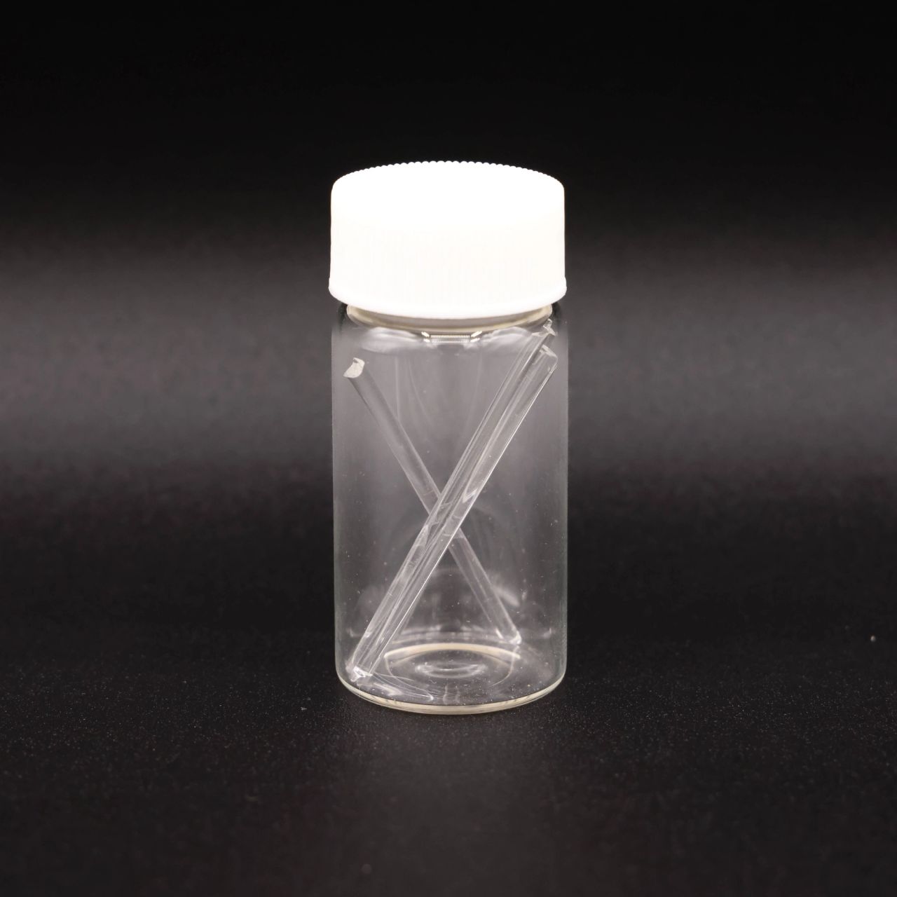 A clear glass vial with a white screw cap holding two fine glass rods crossed inside it.