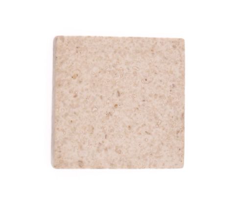 A square tile in pale oatmeal, its surface densely flecked with darker crumbs and pitted with small voids.
