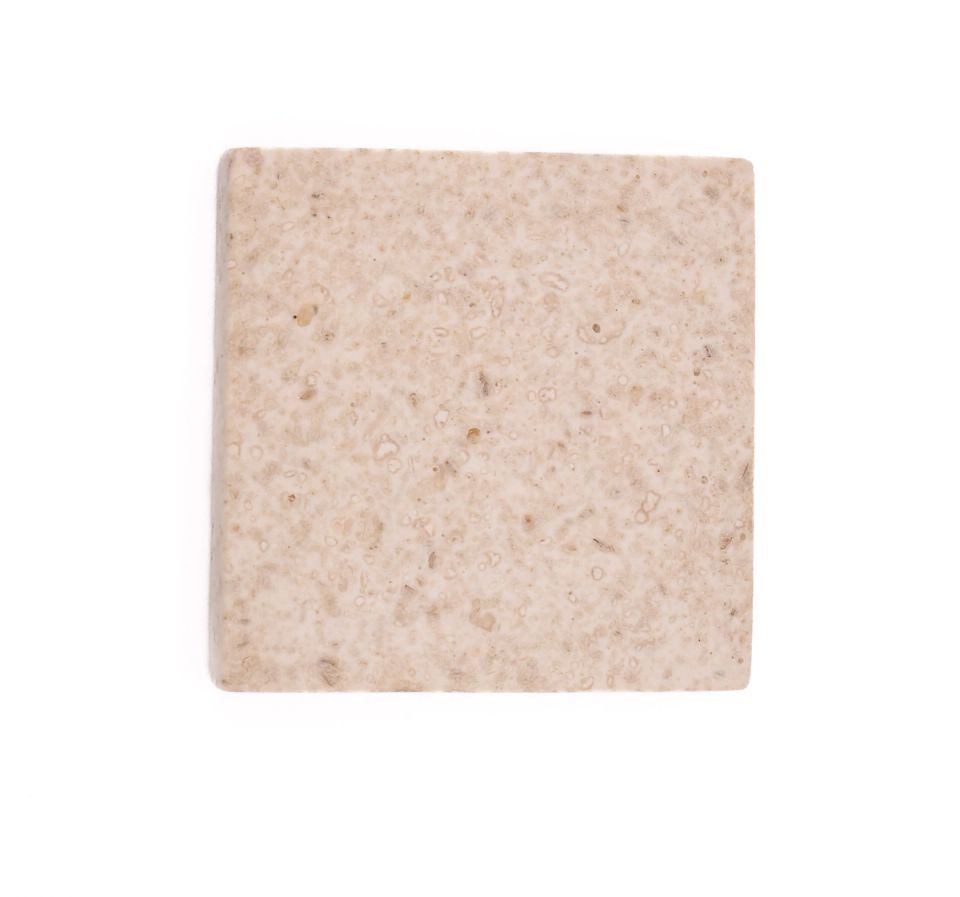 A square tile in pale oatmeal, its surface densely flecked with darker crumbs and pitted with small voids.