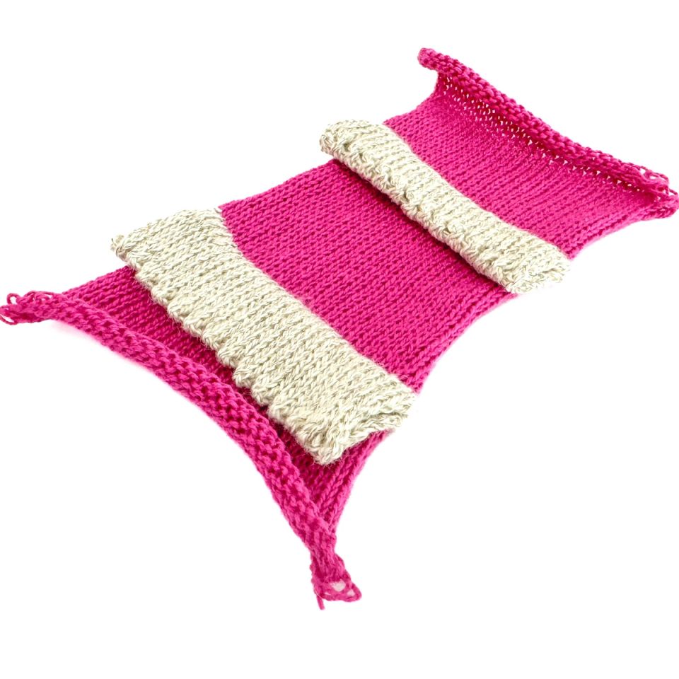 A knitted rectangle in bright magenta crossed by two broad bands of pale cream knitting, its corners curling upward and loose yarn tails trailing from the edges.