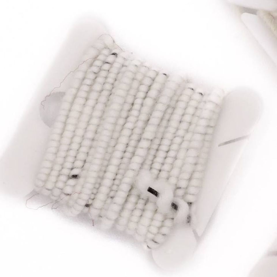 A close view of white beaded cord wound in dense rows on a bobbin, a few dark flecks caught among the coils and a fine dark wire crossing the surface.