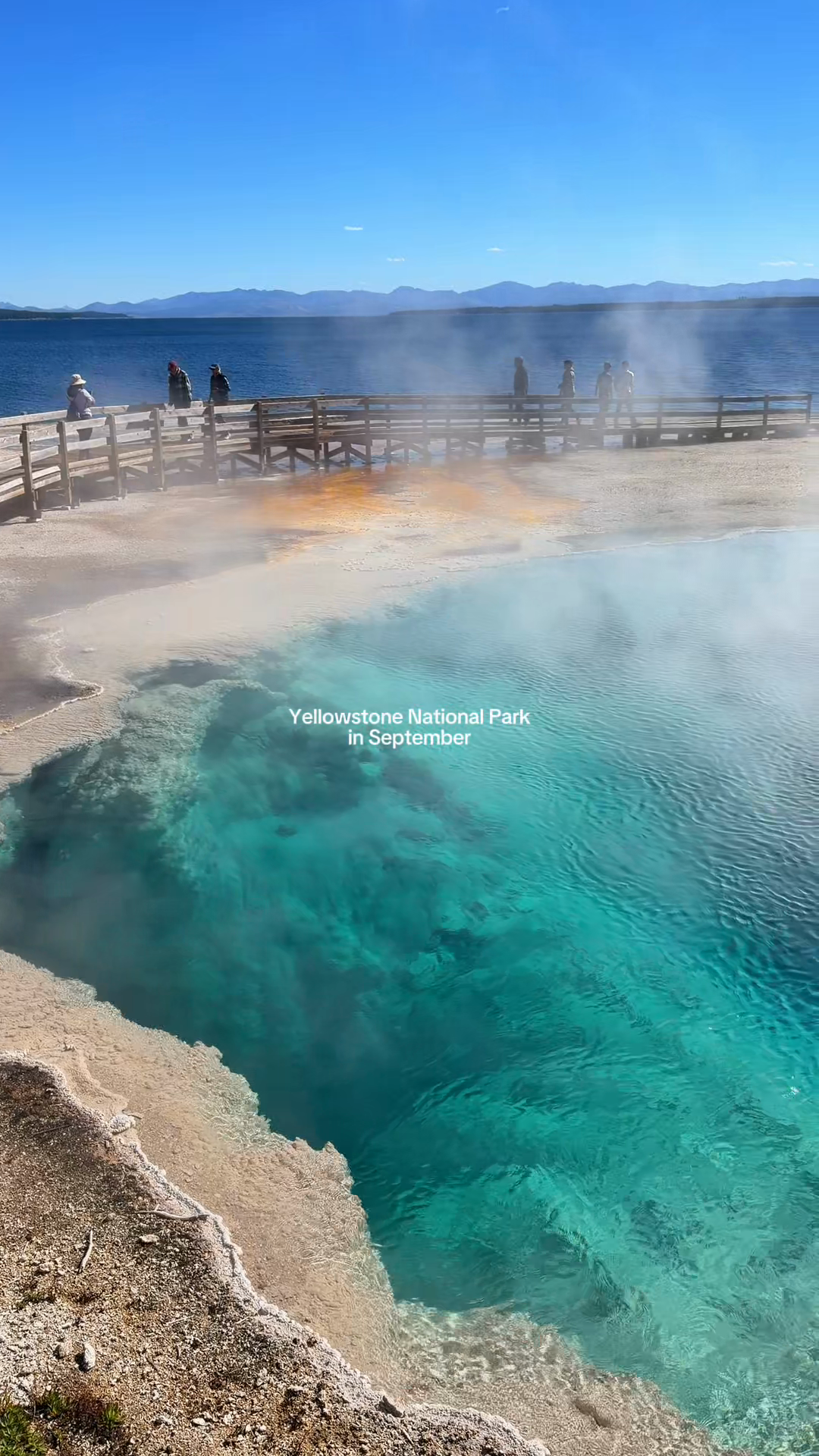 A turquoise thermal pool, pale mineral edge, and distant lake in Yellowstone.