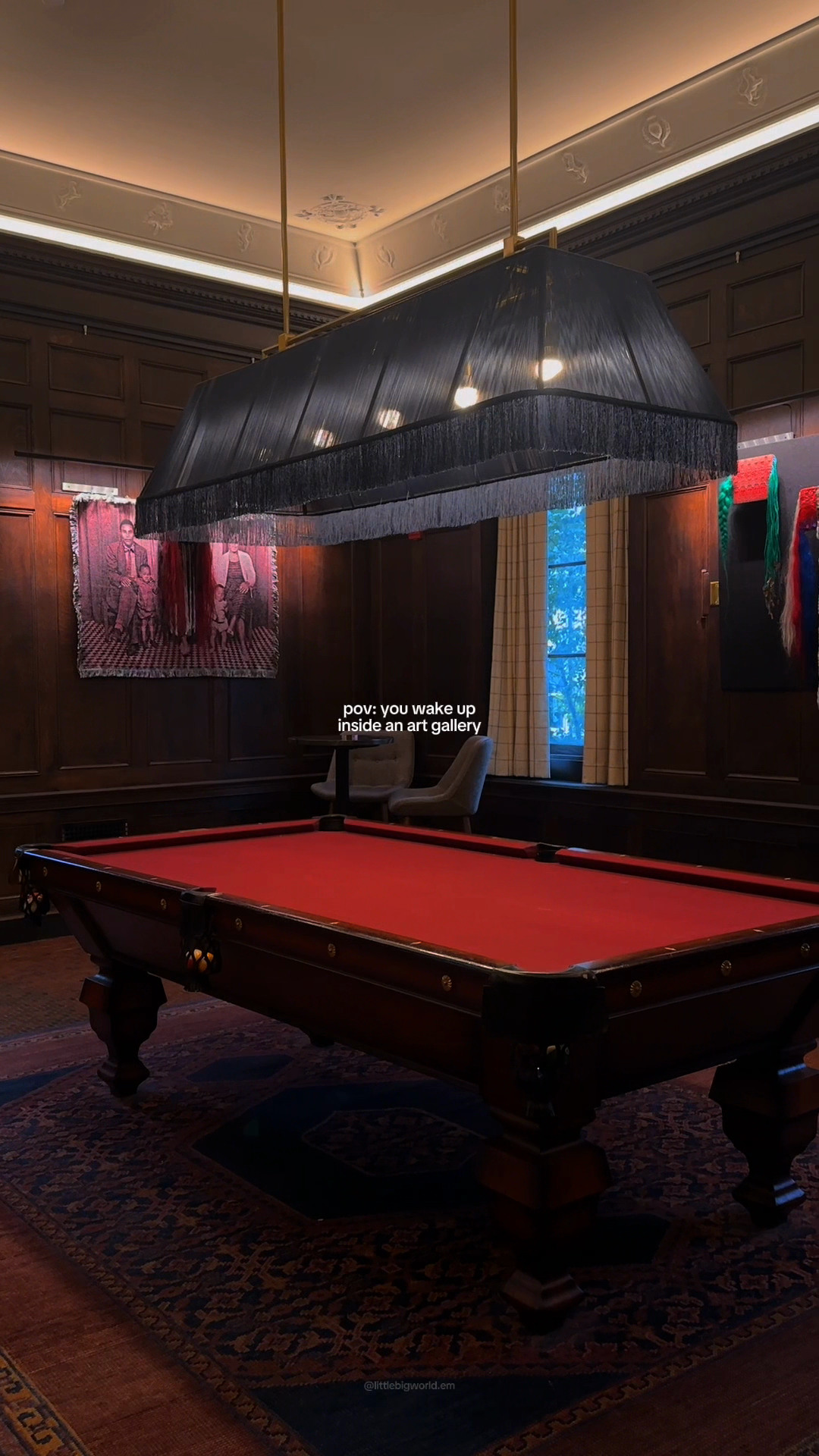 A red-felt billiards table beneath a fringed black lamp in a paneled room with contemporary art.