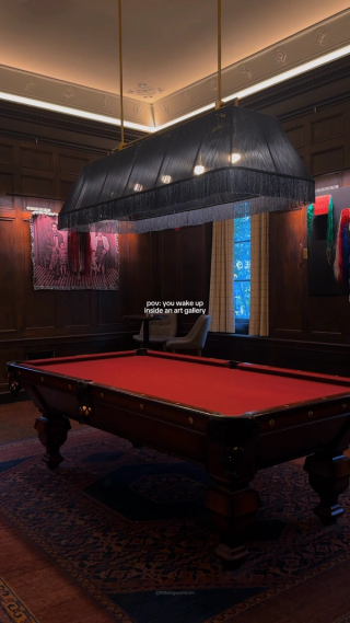 A red-felt billiards table beneath a fringed black lamp in a paneled room with contemporary art.