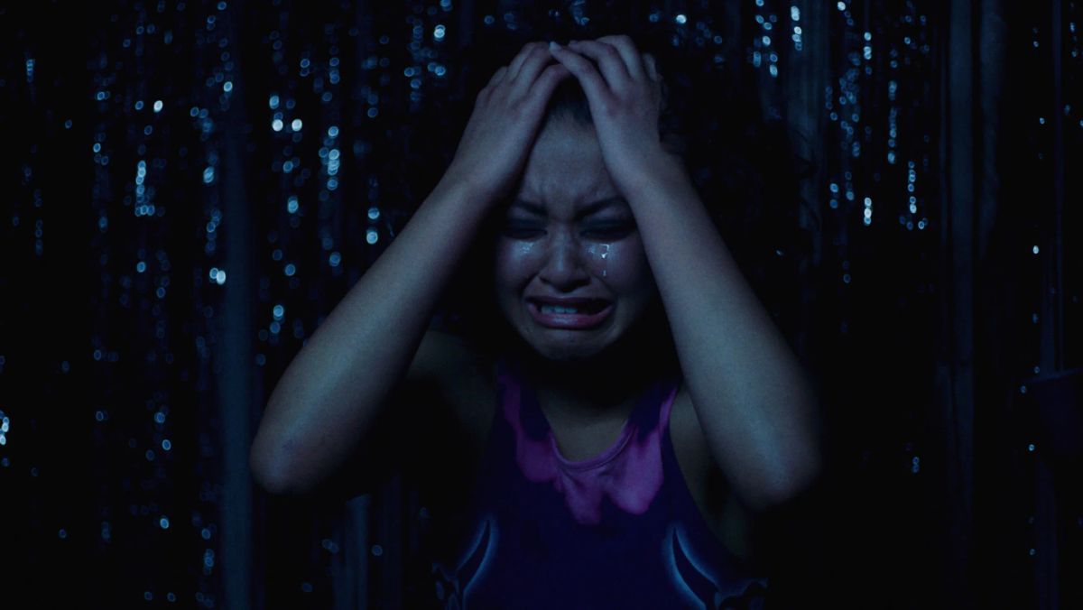A girl cries with her hands in her hair, at night, in front of a glittering curtain.
