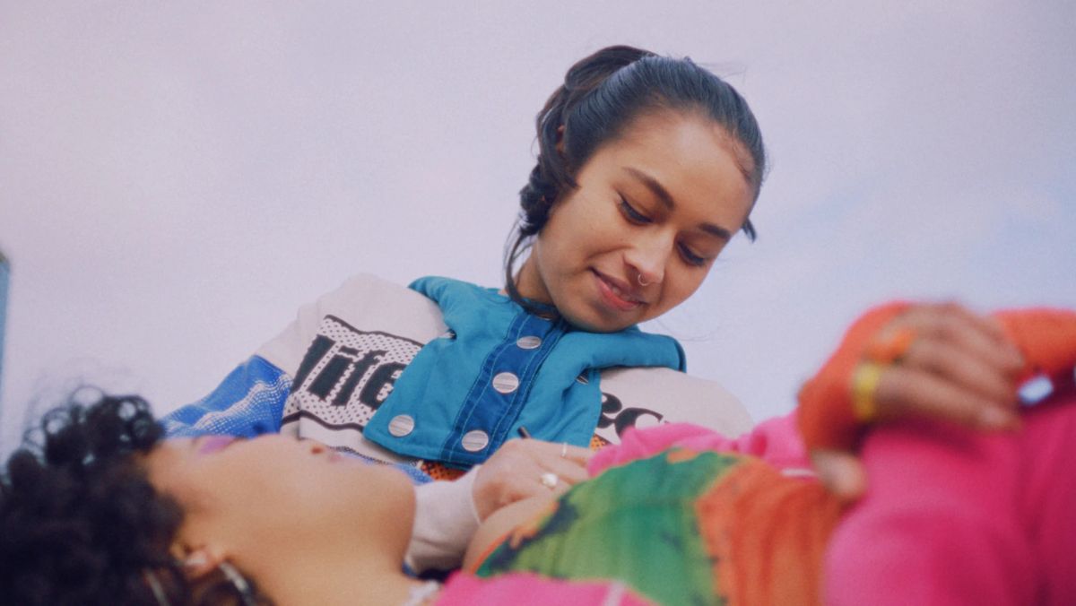 A girl in a blue jacket smiles down at another girl lying on the ground, under a lilac sky.