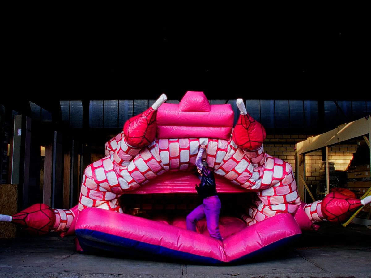 A pink bouncy castle shaped like a monster with an open mouth, in a hangar at night, someone inside the mouth.