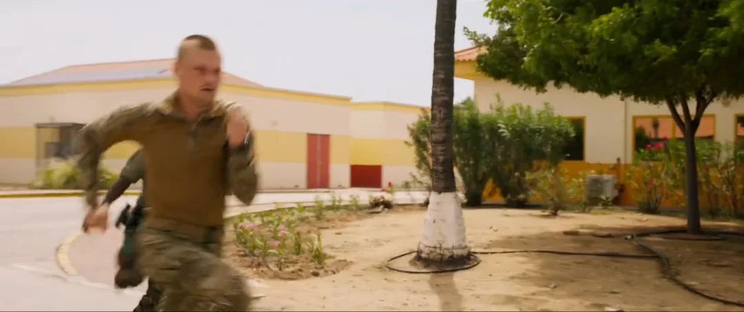 A soldier runs past low yellow buildings and palm trees.