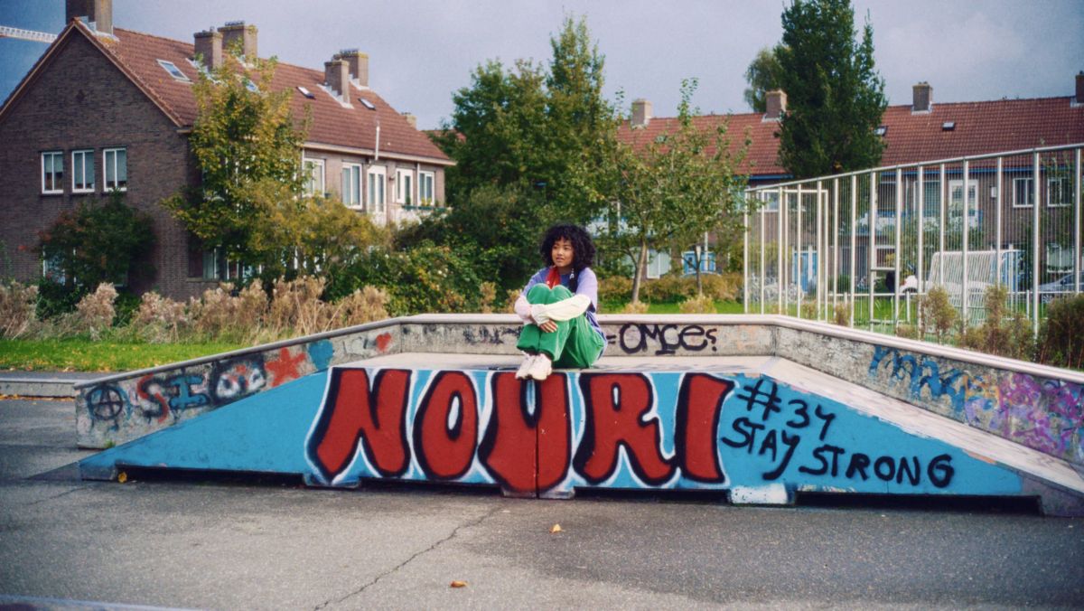 A girl sits on the edge of a graffitied skate bowl in front of a row of brick houses.