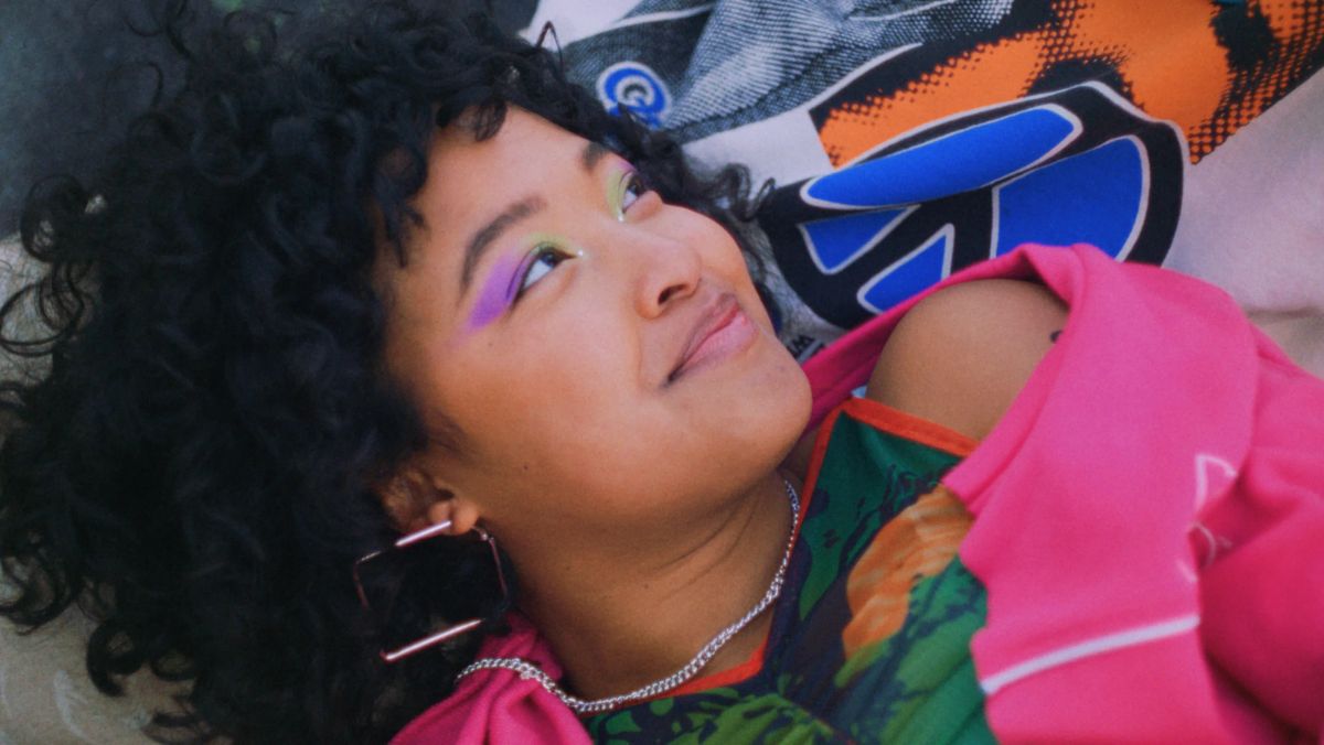 Close-up of a girl with curls and lilac eyeshadow looking up, in a pink jumper, lying on a colourful blanket.