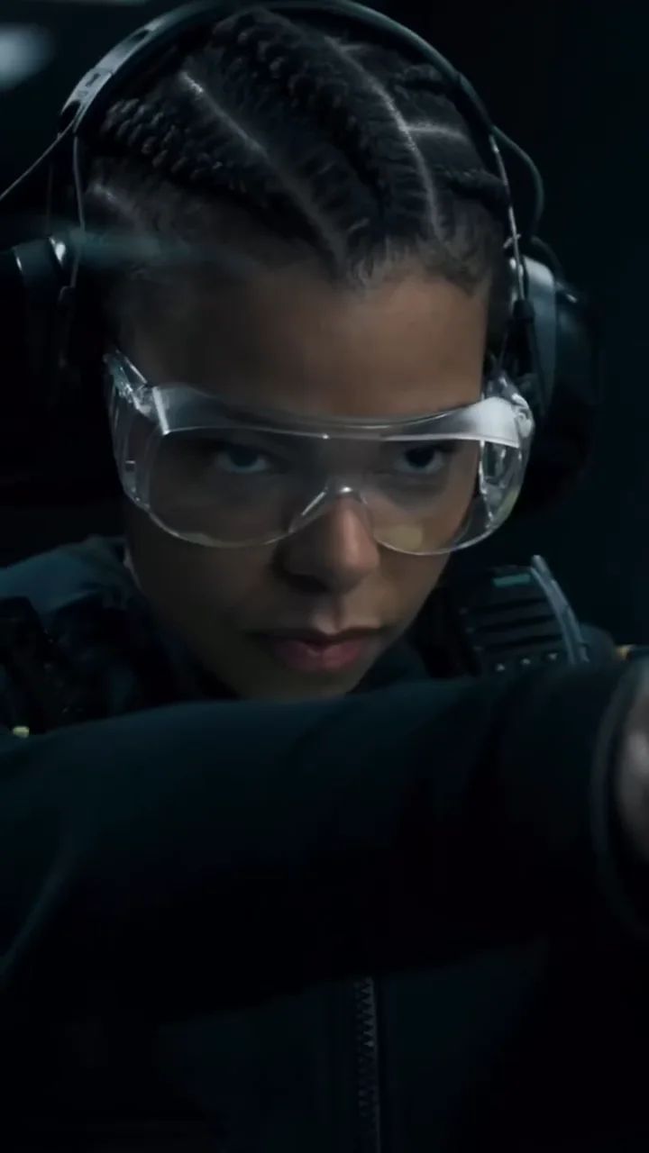 A young woman with ear defenders and safety glasses aims a gun.