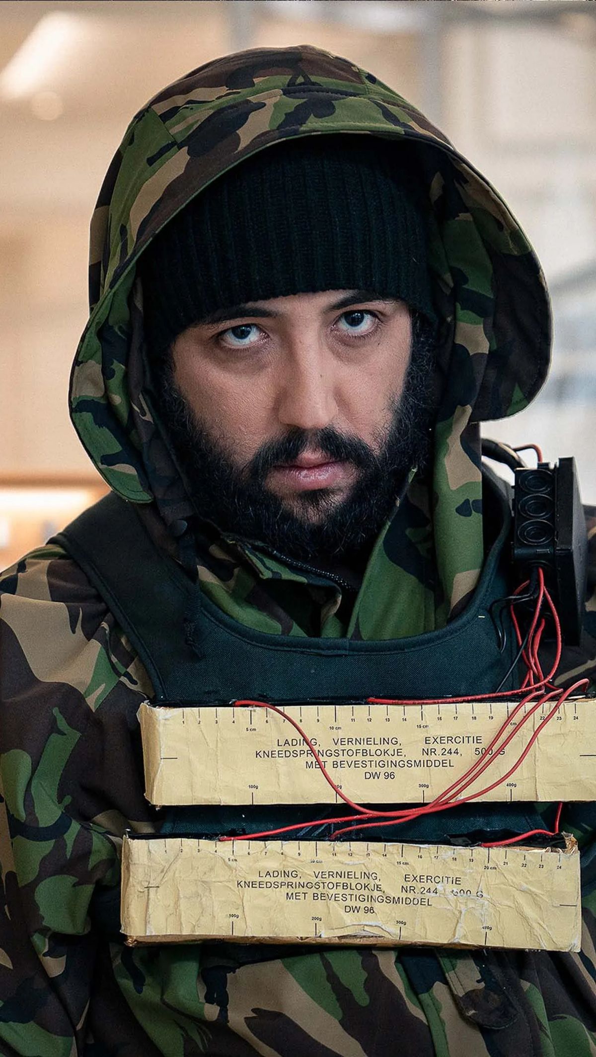 An actor in a camouflage jacket with a hood and a beanie, with fake explosives on his chest.