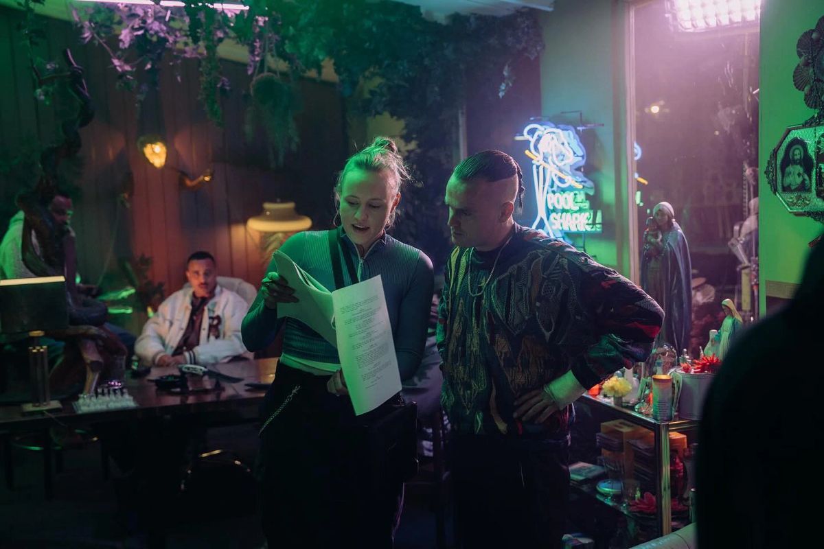 A blonde woman with a script and a man in a colourful jumper talk in neon light.